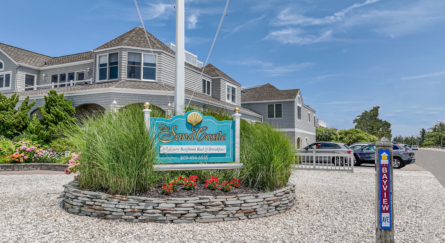 External view of property with light gray shake siding, white trim, green bushes, colorful flower gardens, and sign with property's name