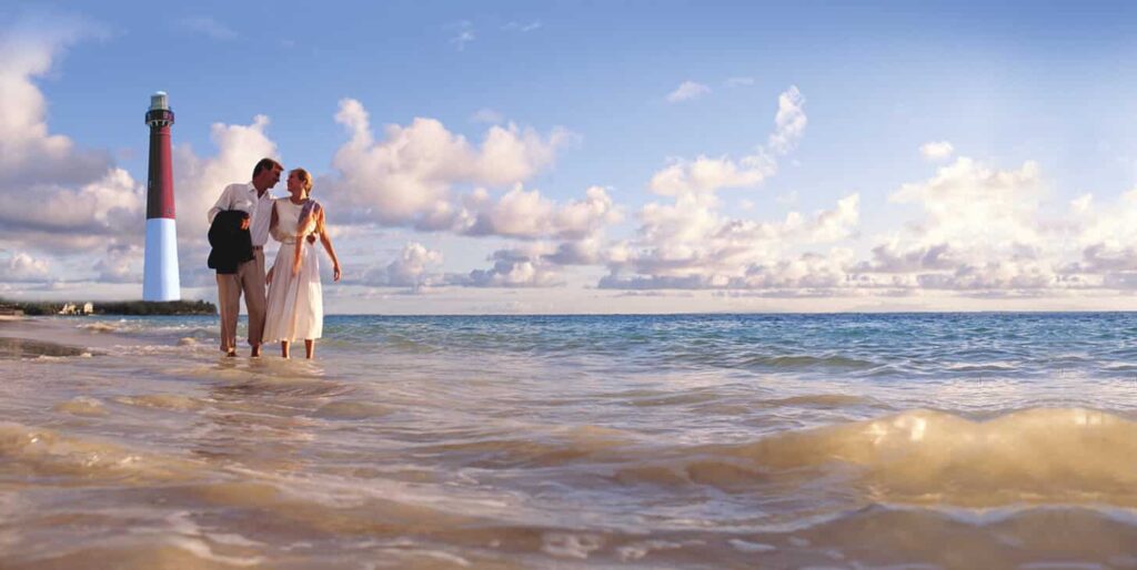 Couple in street clothes strolling in tide, gazing at each other. Lighthouse, sky with puffy clouds, ocean in background.