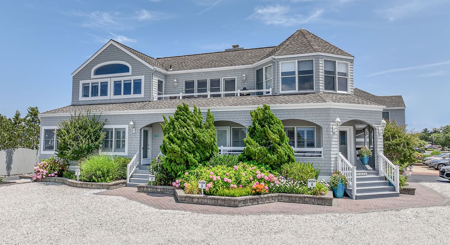 External view of property with light gray shake siding, white trim, green bushes, and colorful flower gardens