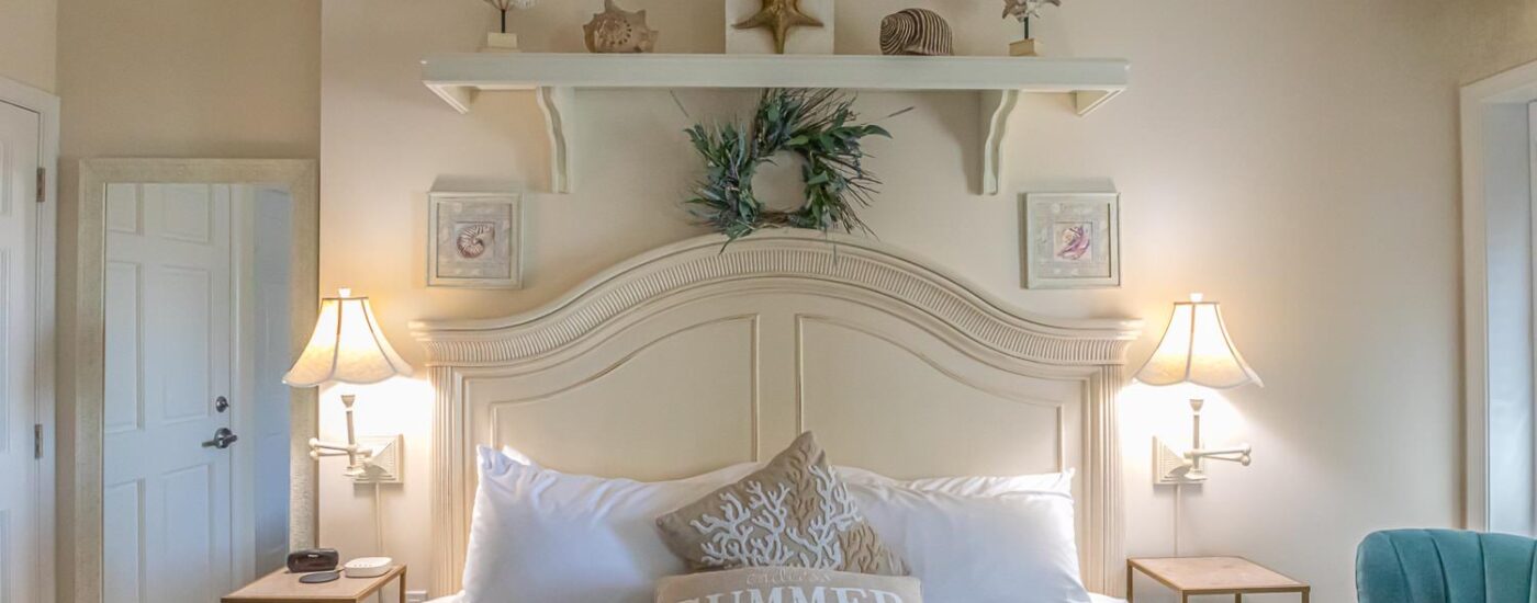 Bedroom with cream walls, cream wood headboard, white and gray bedding, and turquoise upholstered chair