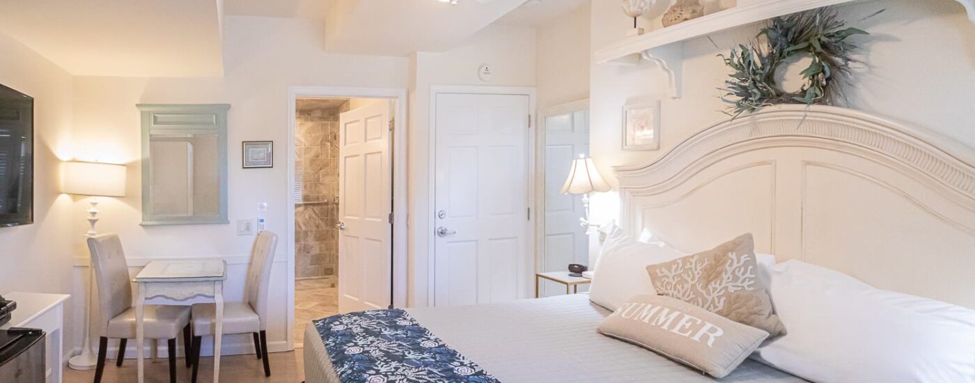 Bedroom with white walls, white wooden headboard, white bedding, sitting area, and view into bathroom