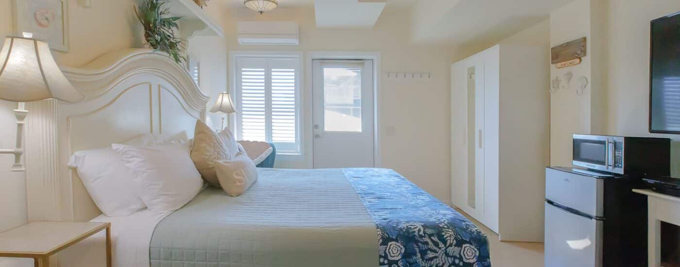 Bedroom with white walls, white wooden headboard, light green and white bedding, white armoire, mini fridge, and microwave