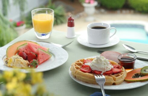 Close up view of a white plate with Belgian waffle covered in strawberries and whipped cream and another white plate with fruit and a scone