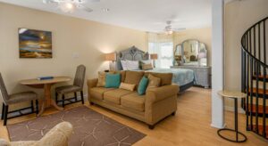 Bedroom suite with cream walls, hardwood flooring, gray wooden bed, multicolored bedding, brown upholstered sofa, sitting area and spiral staircase