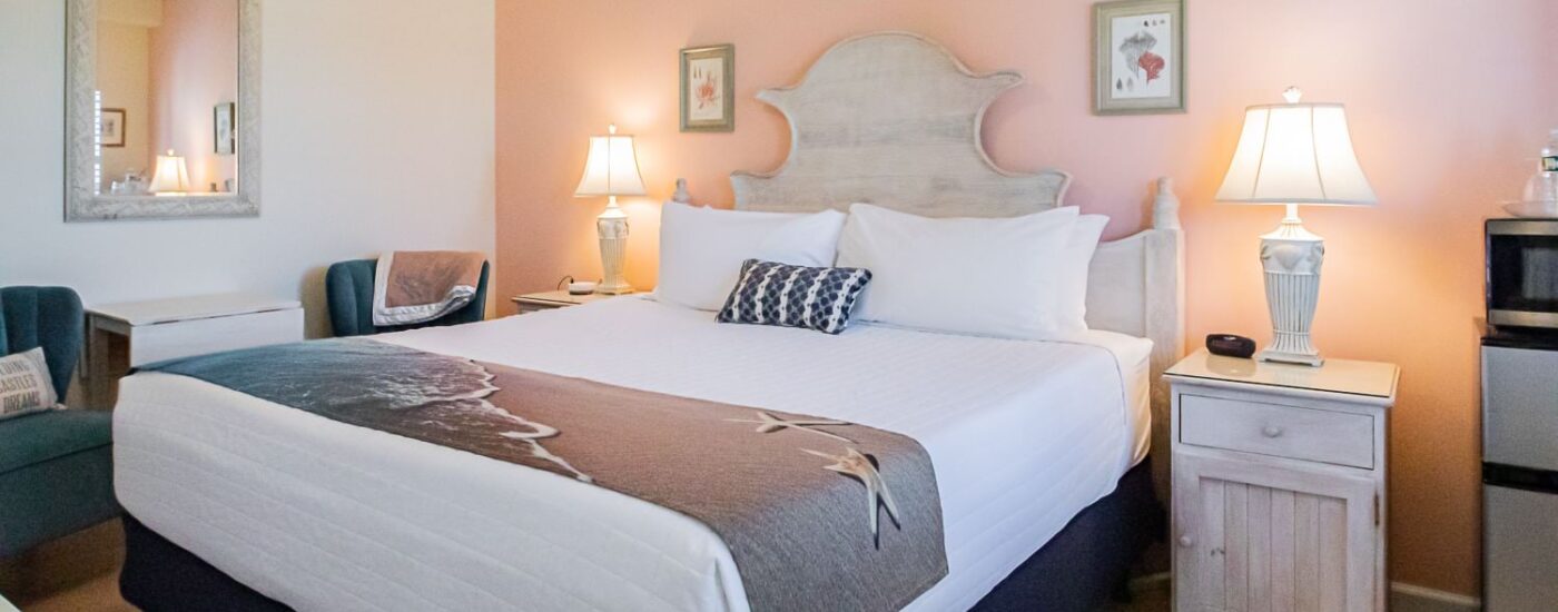 Bedroom with peach and white walls, hardwood flooring, wood bed, white bedding, two nightstands each with a lamp, and sitting area