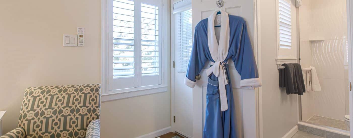Bedroom with light cream walls, hardwood flooring, cream and green upholstered chair, blue and white hanging robe, and view into bathroom