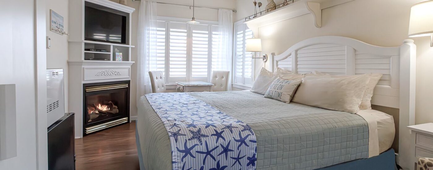 Bedroom with light cream walls, hardwood flooring, white wood bed, light green and white bedding, mini fridge, microwave, and fireplace