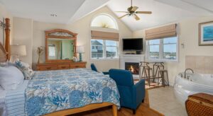 Bedroom suite with cream walls, hardwood flooring, wood bed, multicolored bedding, wood dresser, sitting area, fireplace, and jetted tub