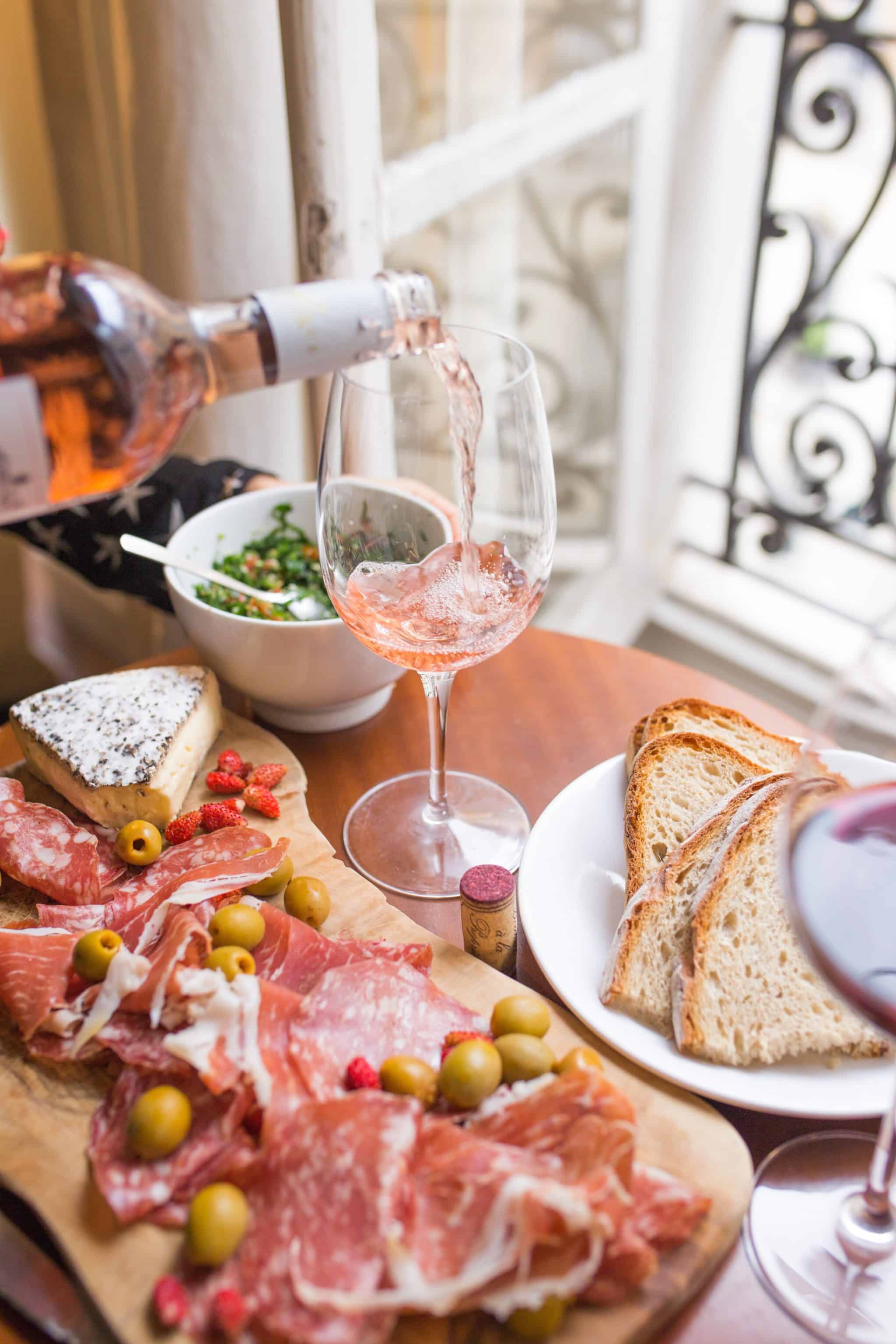 Dining table topped with bread prosciutto, cheese, salad and rosé wine