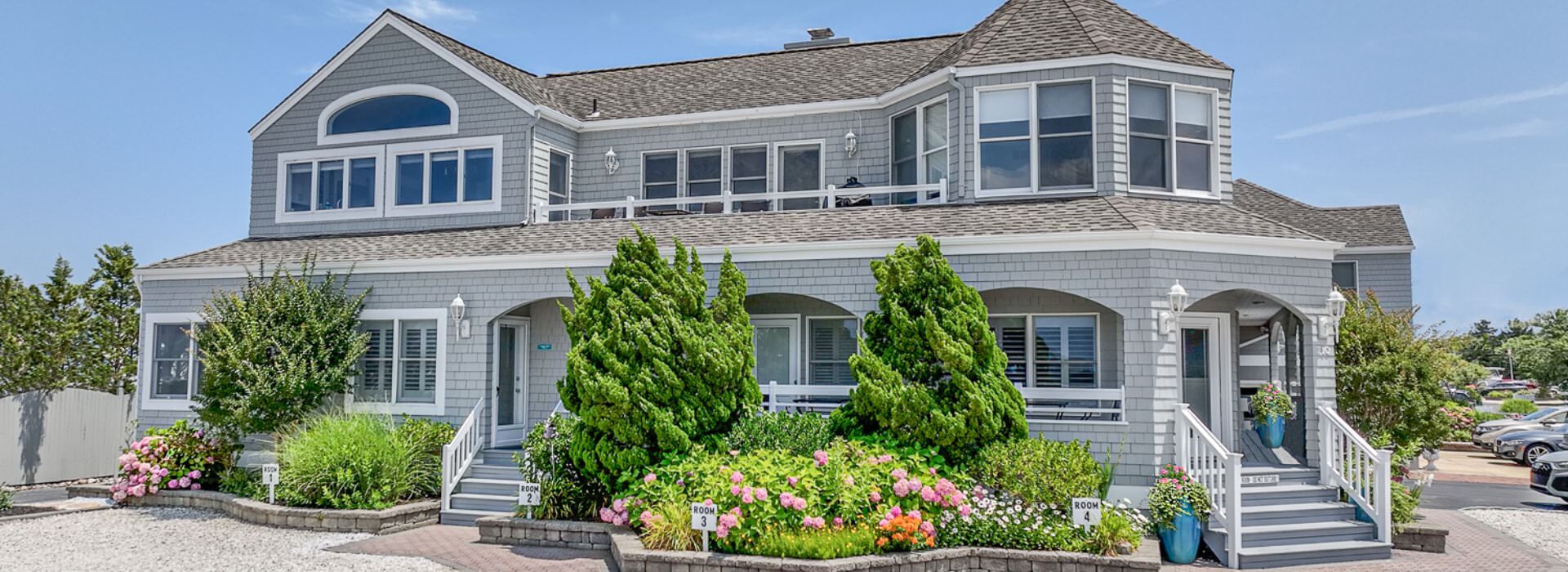 External view of property with light gray shake siding, white trim, green bushes, and colorful flower gardens