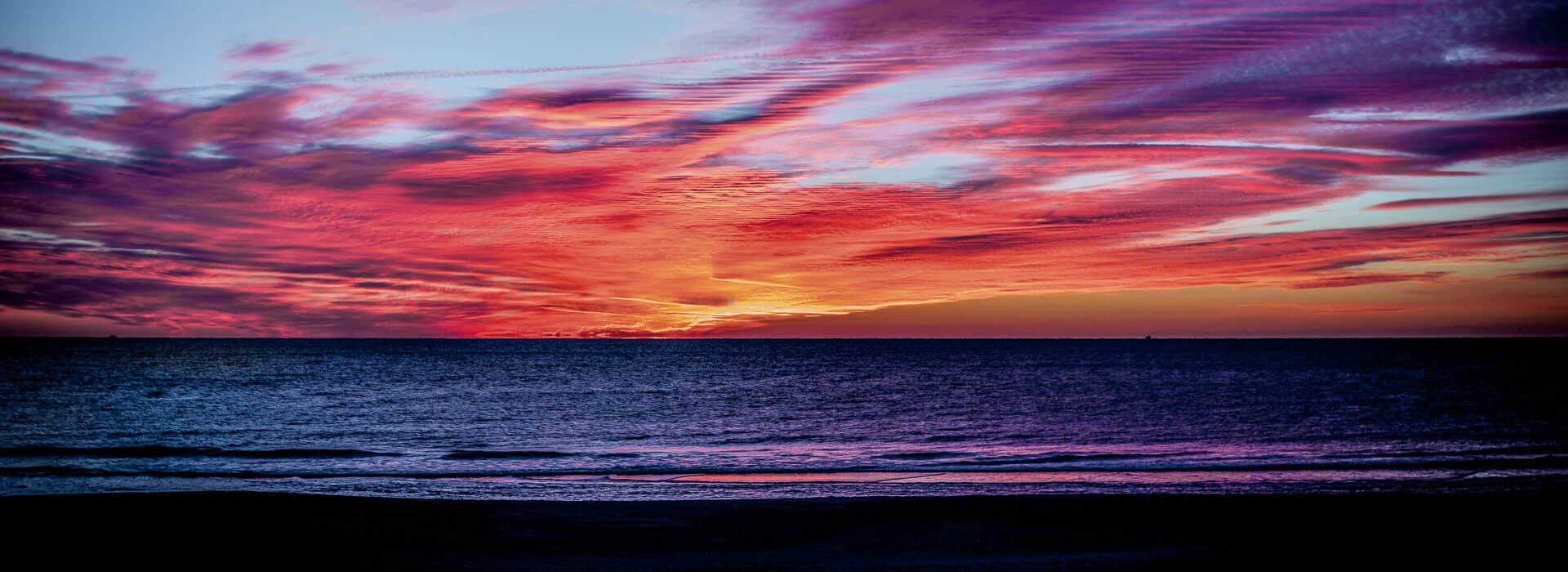 View of a beach with soft rolling waves at dusk with bright orange, pink, blue, and purple clouds in the sky