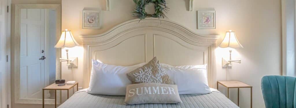 Bedroom with cream walls, cream wood headboard, white and gray bedding, and turquoise upholstered chair