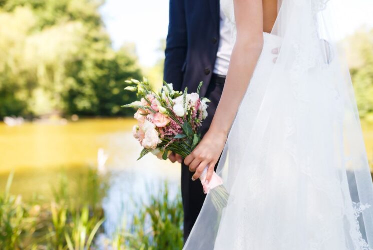 Woman in white dress holding a bouquet of flowers standing by a man in navy suit with water and green vegetation in the background