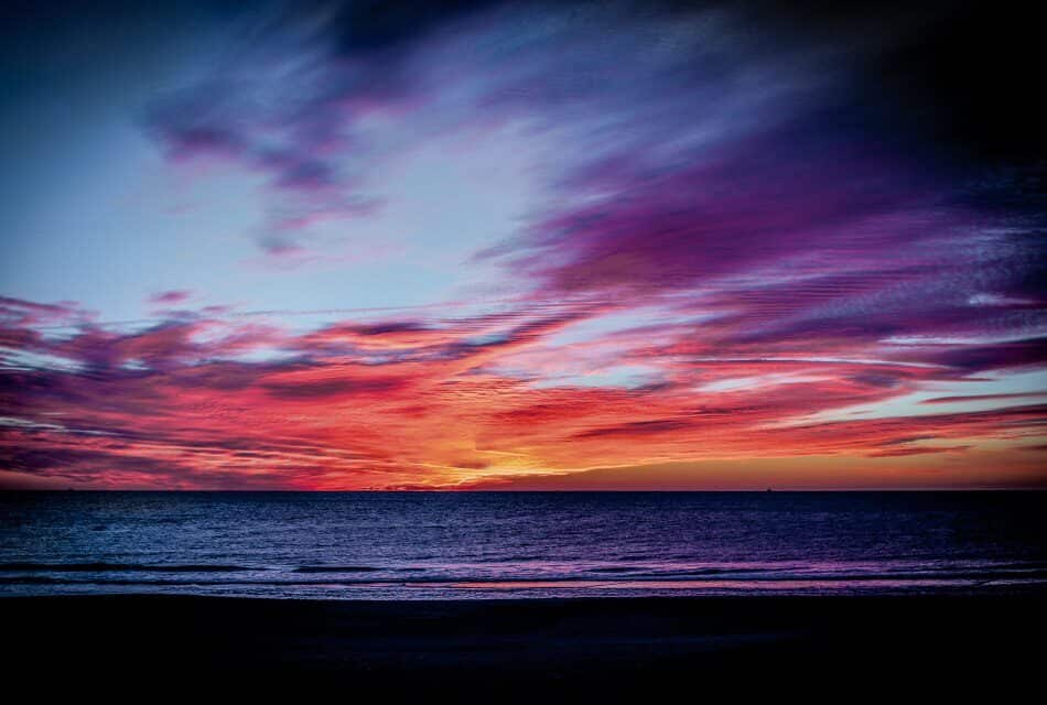 View of a beach with soft rolling waves at dusk with bright orange, pink, blue, and purple clouds in the sky